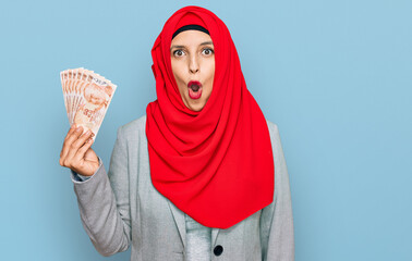 Sticker - Beautiful hispanic woman wearing islamic hijab holding turkish lira banknotes scared and amazed with open mouth for surprise, disbelief face