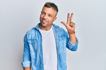 Wall Mural - Handsome muscle man wearing casual denim jacket showing and pointing up with fingers number three while smiling confident and happy.