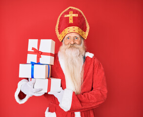 Wall Mural - Old senior man with grey hair and long beard wearing saint nicholas costume holding presents looking positive and happy standing and smiling with a confident smile showing teeth