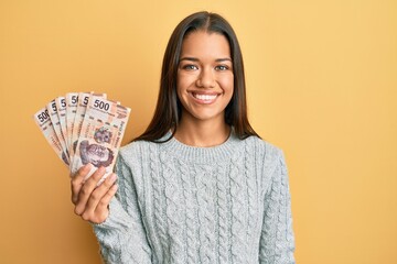 Canvas Print - Beautiful hispanic woman holding 500 mexican pesos banknotes looking positive and happy standing and smiling with a confident smile showing teeth