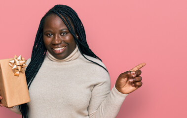 Sticker - Young black woman with braids holding gift smiling happy pointing with hand and finger to the side