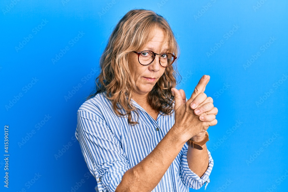 Middle age blonde woman wearing casual clothes and glasses holding symbolic gun with hand gesture, playing killing shooting weapons, angry face