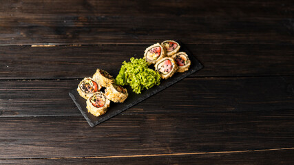 Wall Mural - Custom sushi roll in tempura with nori, fresh salmon, tuna, avocado, masago caviar, drizzled with pineapple sauce with salad pouring as decoration on a black plate on a wooden table and background.