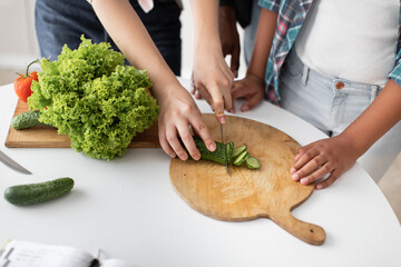 Close up cropped top view of hands of mother and mixed race daughter chopping cucumber on chopping board