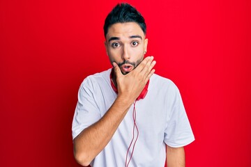 Young man with beard listening to music using headphones looking fascinated with disbelief, surprise and amazed expression with hands on chin