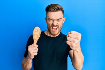 Wall Mural - Young redhead man holding hairbrush annoyed and frustrated shouting with anger, yelling crazy with anger and hand raised