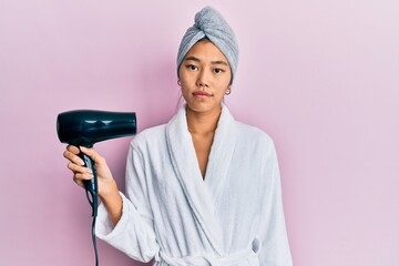 Poster - Young chinese woman wearing shower towel cap and bathrobe holding dryer thinking attitude and sober expression looking self confident