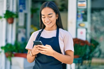 Poster - Young latin shopkeeper girl smiling happy using smartphone at florist.