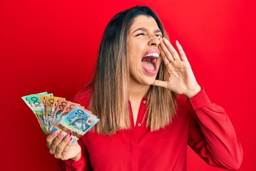 Poster - Beautiful brunette woman holding australian dollars shouting and screaming loud to side with hand on mouth. communication concept.