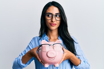 Wall Mural - Beautiful hispanic woman holding piggy bank with glasses smiling looking to the side and staring away thinking.