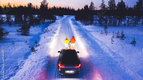 Aerial back view of romantic couple of tourists standing near car while headlights lighting them way on in winter darkness, bird\'s eye view of vehicle in snowy north lands. Wanderlust in road trip