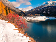 Autumn In The Mountains With Some Snow On The Grounds And Red And Yellow Bushes And Trees Around A Blue Blank Lake Between The Hills And Mountain Peaks