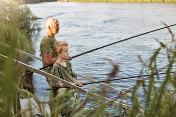 Wall Mural - European grey haired mature father with son outdoors fishing by lake or river, standing near water with fishing rods in hands, dress casually, enjoying hobby and nature.