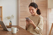 © Seventyfour - Warm toned portrait of smiling young woman using smartphone at home office while enjoying remote work in cozy interior, copy space