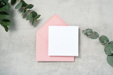 Real photo. Pink envelope square invitation card mockup with a eucalyptus branch. Top view with copy space, light gray concrete background. Template for branding and advertising