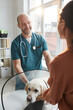 © Seventyfour - Vertical portrait of mature veterinarian examining dog at vet clinic while talking to young woman