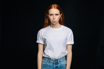 Wall Mural - Indecisive young woman wearing T-shirt and denim biting lips apologizing on isolated back background, looking away . Pretty redhead lady model emotionally showing facial expressions.