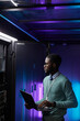 © Seventyfour - Vertical portrait of young African American data engineer holding laptop while working with supercomputer in server room lit by blue light, copy space