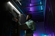 © Seventyfour - Low angle portrait of young African American data engineer working with supercomputer in server room lit by blue light and holding laptop, copy space
