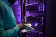 © Seventyfour - Cropped shot of African American data engineer holding laptop while working with supercomputer in server room lit by blue light, copy space