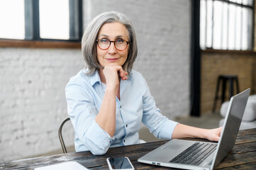 Intelligent mature senior female business owner in glasses with hand on a chin looking at the camera, sitting at the desk in front of laptop. A successful businesswoman enjoying and loving her job