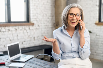 Excited modern senior businesswoman manager in glasses and casual attire standing in the office, taking break to call and talk with family, holding mobile phone, discussing news, gossiping, gesturing