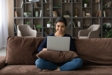 Wall Mural - Happy millennial Indian woman sit relax on couch in living room study distant on computer gadget. Smiling young ethnic female rest on sofa at home office work online on laptop. Technology concept.