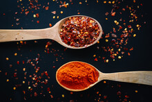 Fresh natural spices: red paprika and red chili pepper in wooden spoons on the black background. Top down view.