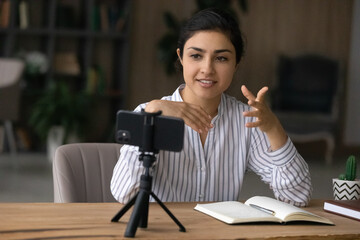 Young Indian woman speaker or coach record live video broadcast on cellphone camera on tripod. Successful ethnic female teacher or trainer talk on webcam, have online call or webinar on cell.