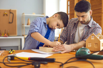 Wall Mural - Adult man teaching kid to make wooden toys. Concentrated little child and his father building house models at home. Dad and son working with wood at carpenter's workshop as part of school DIY project