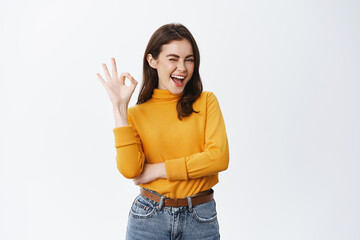 confident young woman winking to assure you, showing okay sign and smiling, guarantee good quality, 