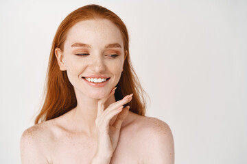 Wall Mural - Skincare. Close-up of redhead woman with pale soft skin, smiling white teeth and touching clean no makeup face, looking aside, standing over white background naked