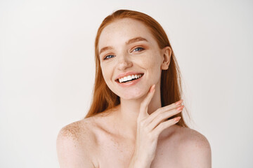 Wall Mural - Beauty. Close-up of young beautiful redhead woman smiling at camera, touching perfect clean skin on face and looking happy, standing naked over white background