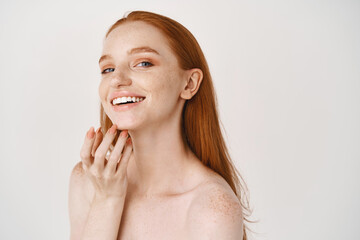 Close-up of smiling redhead woman with pale skin and freckles touching soft, perfect face, using skincare cream, standing over white background