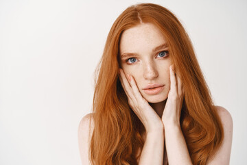 Wall Mural - Beauty and skincare. Close-up of redhead pale woman touching face, looking at camera sensual, showing perfect no makeup skin, standing over white background