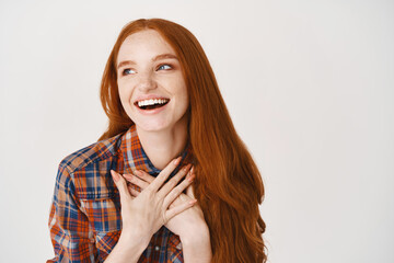 Wall Mural - Close-up of happy ginger girl with pale skin and blue eyes smiling thankful, expressing gratitude and joy, holding hands on heart, standing over white background