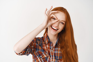 Wall Mural - Close-up of beautiful redhead woman smiling with white teeth, showing okay sign and looking happy, standing over white background