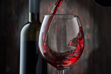 Red wine is poured into a glass from a bottle on a blurred wooden background, a stream of red wine from the bottle swirls in the glass, close-up. Free space for text.