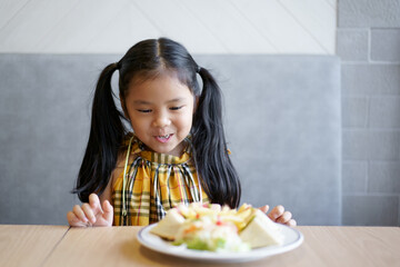 Wall Mural - Asian child hungry or kid girl smile to enjoy happy eating bread or sandwich and vegetable salad on dish and wood table at school or home kitchen and restaurant for breakfast food or snack at lunch