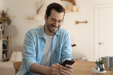 Wall Mural - Happy young Caucasian man sit at desk at home look at smartphone screen texting or messaging online on gadget. Smiling millennial male use cellphone, browse wireless internet. Mobile provider concept.