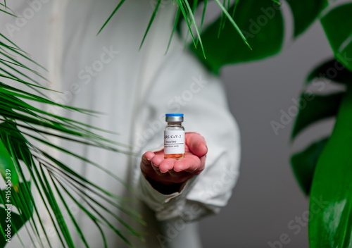 Woman in white shirt show vaccine in a bottle near palm leaves. © Masson
