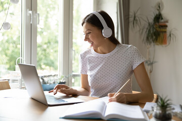 Wall Mural - Happy millennial Caucasian female student in headphones look at laptop screen study distant from home. Smiling young woman in earphones make notes watch webinar on computer, take web course.