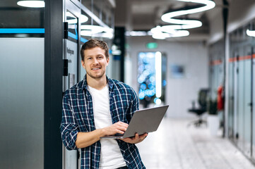 Caucasian guy freelancer with laptop. A successful guy dressed in stylish casual wear stands in a creative office, holds a laptop, looks and smiles friendly at the camera