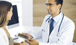 © Iryna - Doctor and patient discussing medical exam results while sitting at the desk in clinic. Male physician using tablet computer for filling up medication history record of young woman. Data in medicine