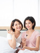 © MergeIdea - Asian two blogger friend show and review beauty cosmetic product including cheek brush, baby pink lipstick and eye shadow palette smile portrait vertical style on bed in white bedroom
