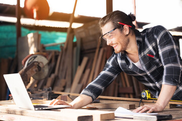 Young woodworker with beard leaning over workbench in his large workshop full of carpentry equipment working online with laptop