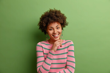 Wall Mural - Portrait of good looking curly haired woman keeps hand under chin smiles broadly has pleased face expression dressed in casual striped jumper isolated over green background. Positive human emotions