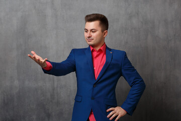 A young business man in a casual blue suit shows a gesture of outstretched hand with an open palm on a gray background.