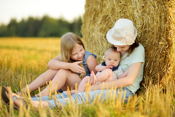 Wall Mural - Big sisters and their baby brother having fun in a wheat field on a summer day. Children playing at hay bale field during harvest time. Kids enjoying warm sunset outdoors.