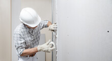 Man Drywall Worker Or Plasterer Putting Mesh Tape For Plasterboard On A Wall Using A Spatula And Plaster. Wearing White Hardhat, Work Gloves And Safety Glasses. Image With Copy Space.